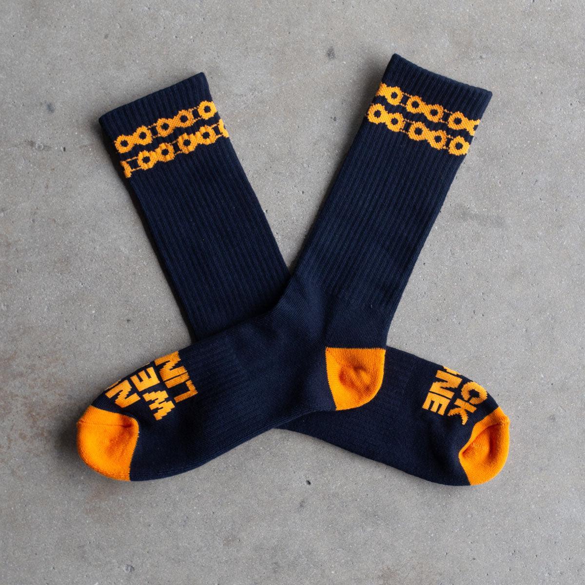 Backbone No Weak Links Socks: Black socks with orange toes and heels, chain-link pattern by the cuffs, and bold orange "NO WEAK LINKS" text on the soles—laid out on concrete to show strength in every step.