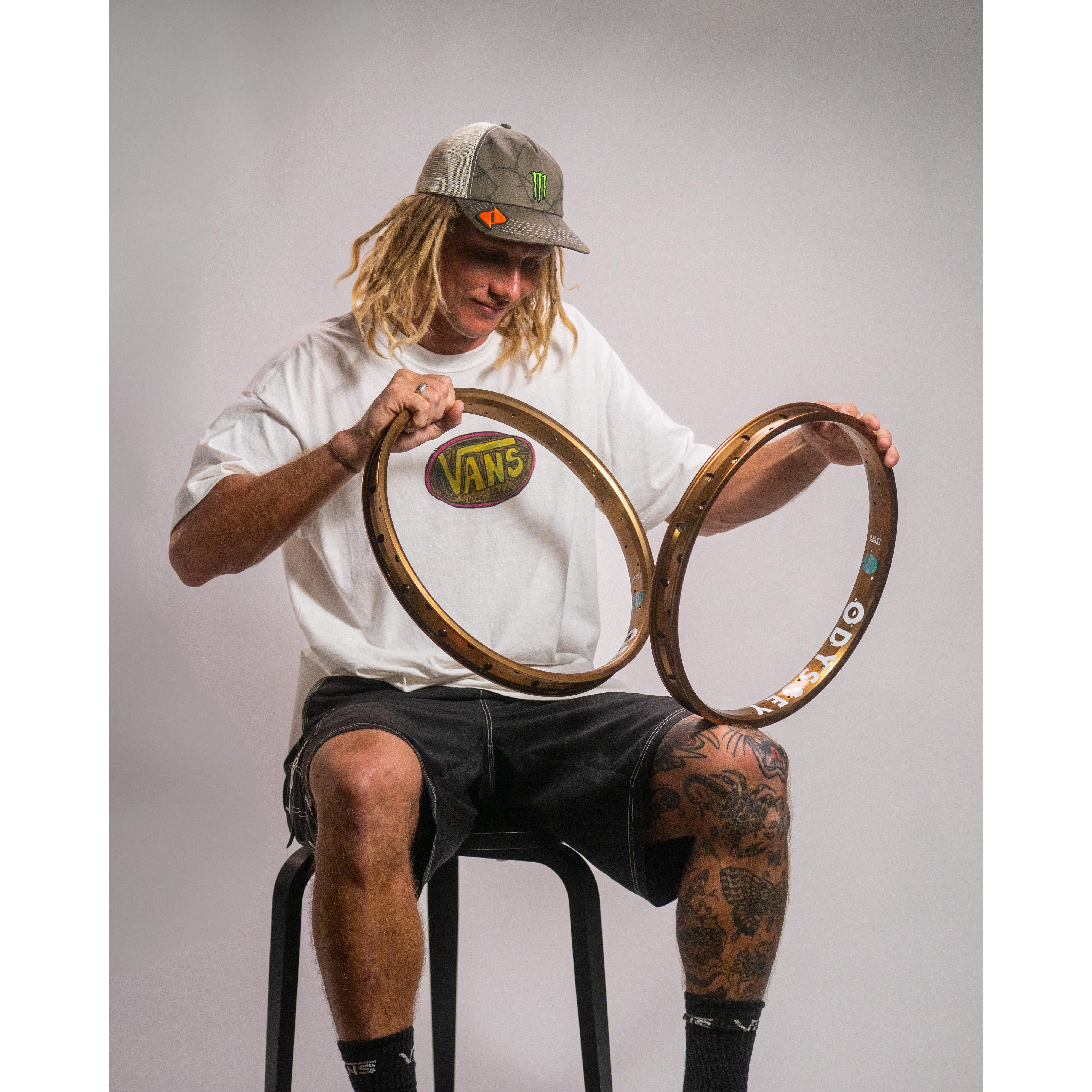 A person with long dreadlocks in a Vans t-shirt and cap sits on a stool, holding two Odyssey Big Boyd Rims—HARD ANODIZED BLACK with extra-wide cross-sections—set against a plain backdrop.