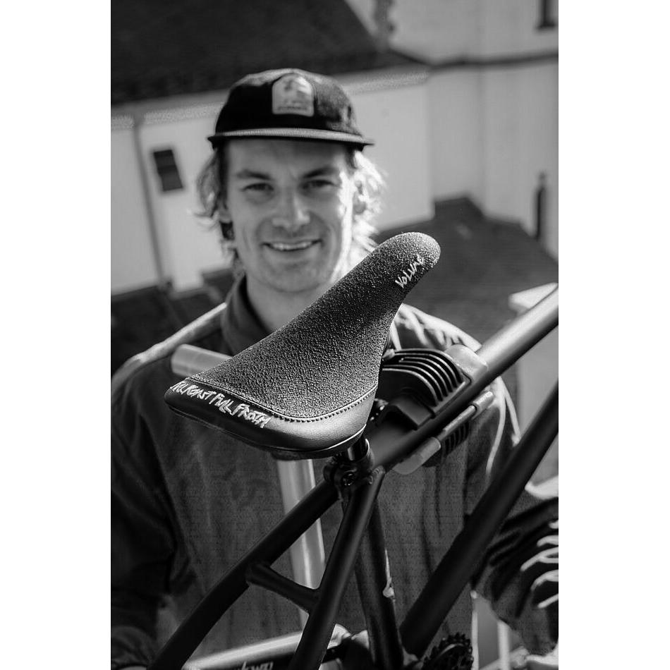 A man in a cap smiles outdoors behind a close-up of the Volume Full Roast Full Froth railed bicycle seat by Paul Theolen, attached to a bike frame.