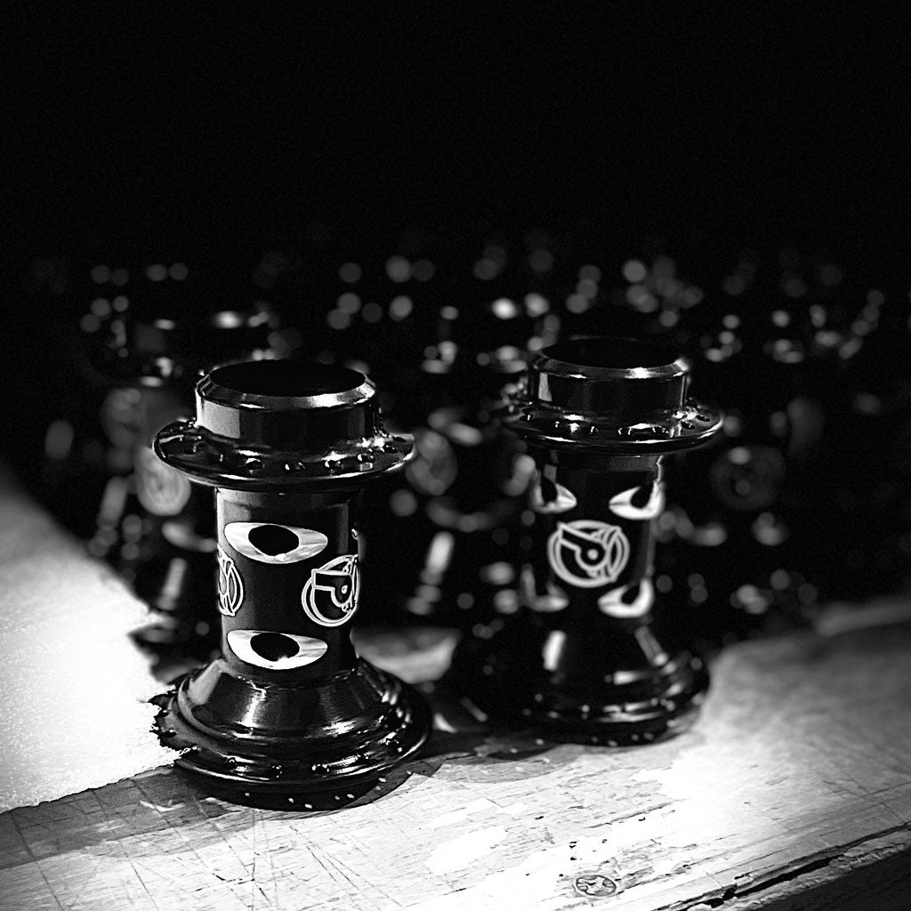 Black and white photo of several identical mechanical parts lined up on a surface, featuring cylindrical shapes with detailed patterns and designs, resembling the precision of Profile Racing's Limited Edition Cats Eye Hub Set.