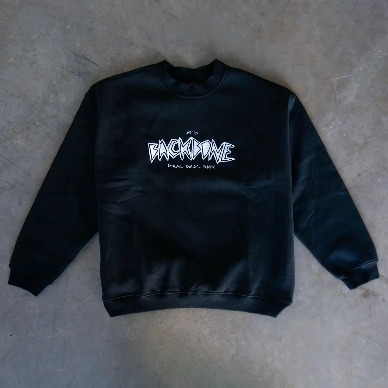 The Backbone Real Deal Crewneck features "BACKBONE REAL DEAL BMX" in white graffiti-style text on the front, laid flat on a gray surface—an ideal winter essential.