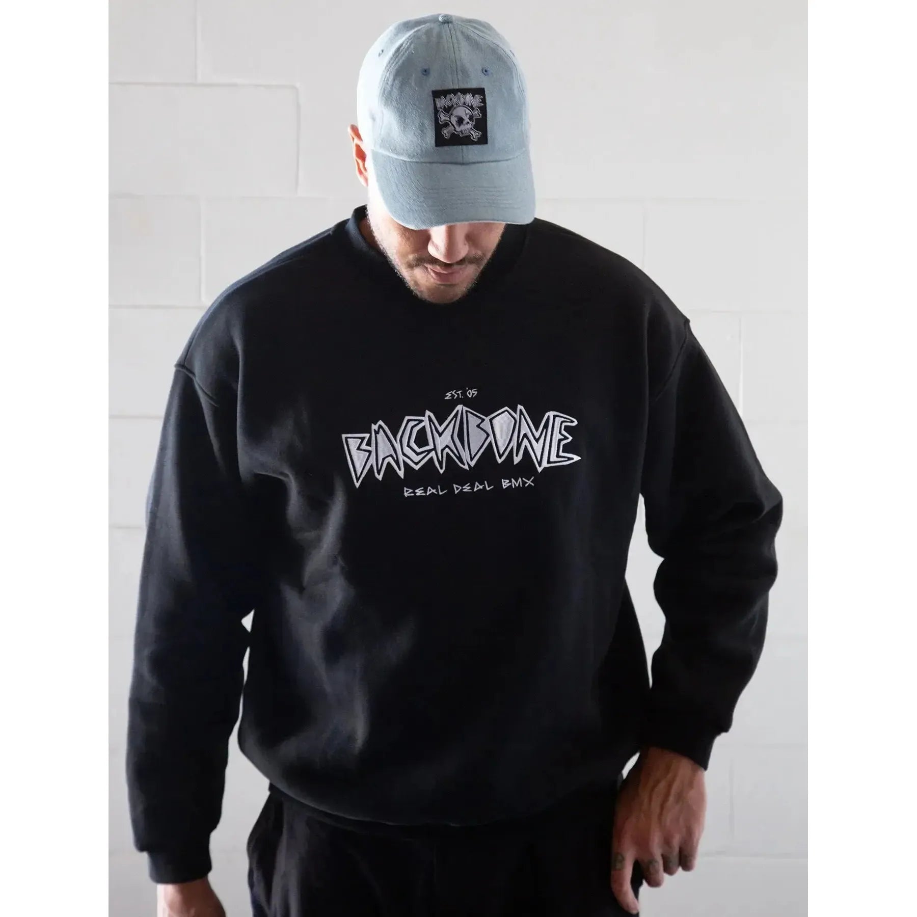 A person in a light blue cap and the Backbone Real Deal Crewneck with "REAL DEAL BMX" on the front stands against a white brick wall, looking down—ideal for your winter kit.