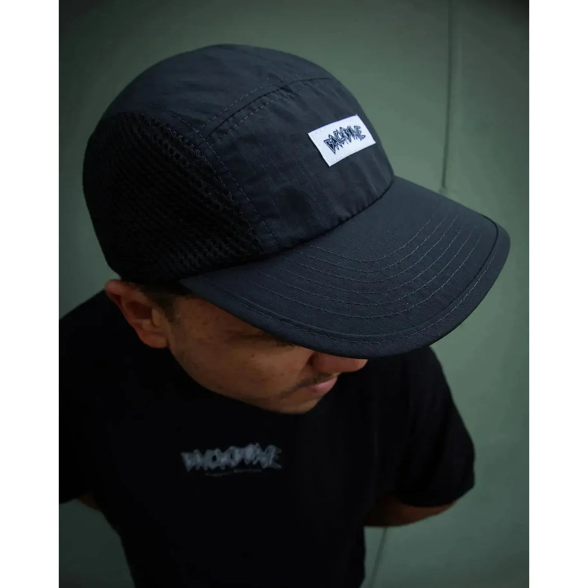 Wearing a black Backbone Ride Club Hat featuring a “BADWAVE” patch, along with a matching black “BADWAVE” t-shirt, the person stands against a plain greenish wall.
