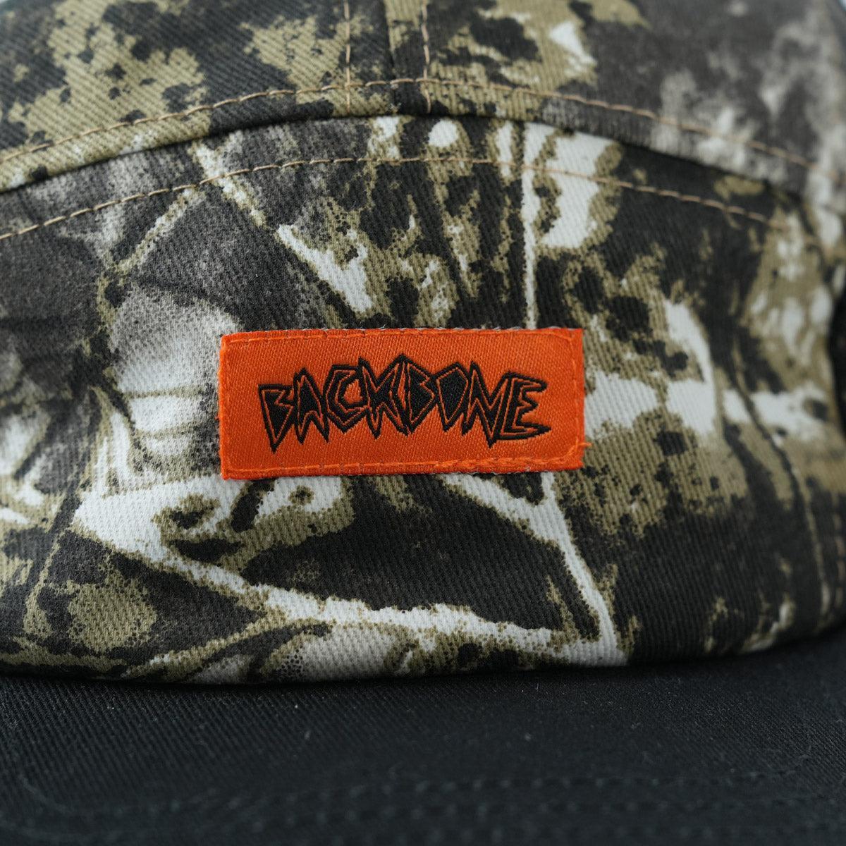 A close-up of the Backbone Ride Club Hat, a camouflage-patterned 5 panel cap featuring an orange patch with "BACKBONE" in bold, stylized lettering.