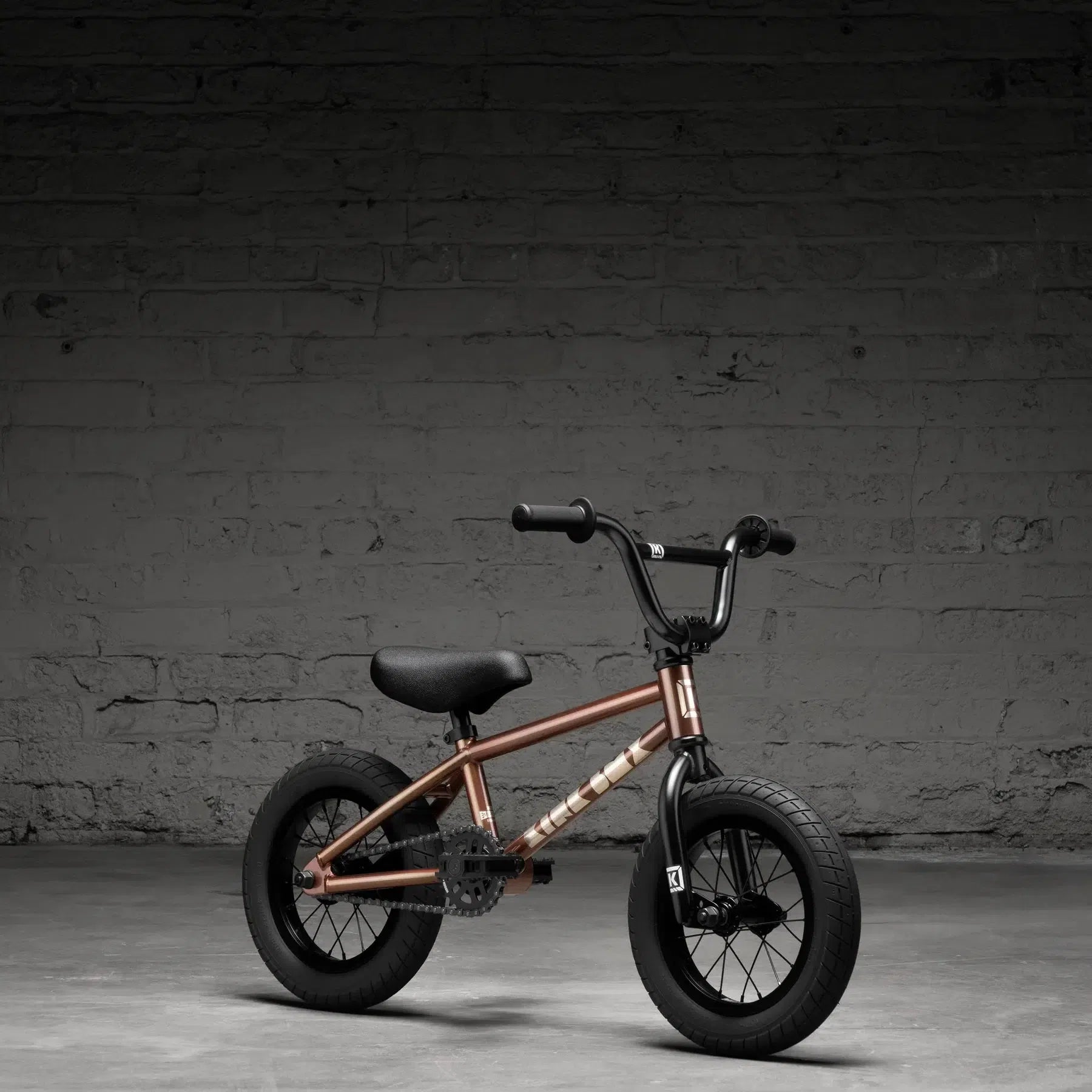 The Kink Roaster 12 Inch Bike (2026), ideal as a child's first BMX, boasts a bronze frame, black seat and wheels. It's set against a dark brick wall background and includes a reliable coaster brake for safe stopping.
