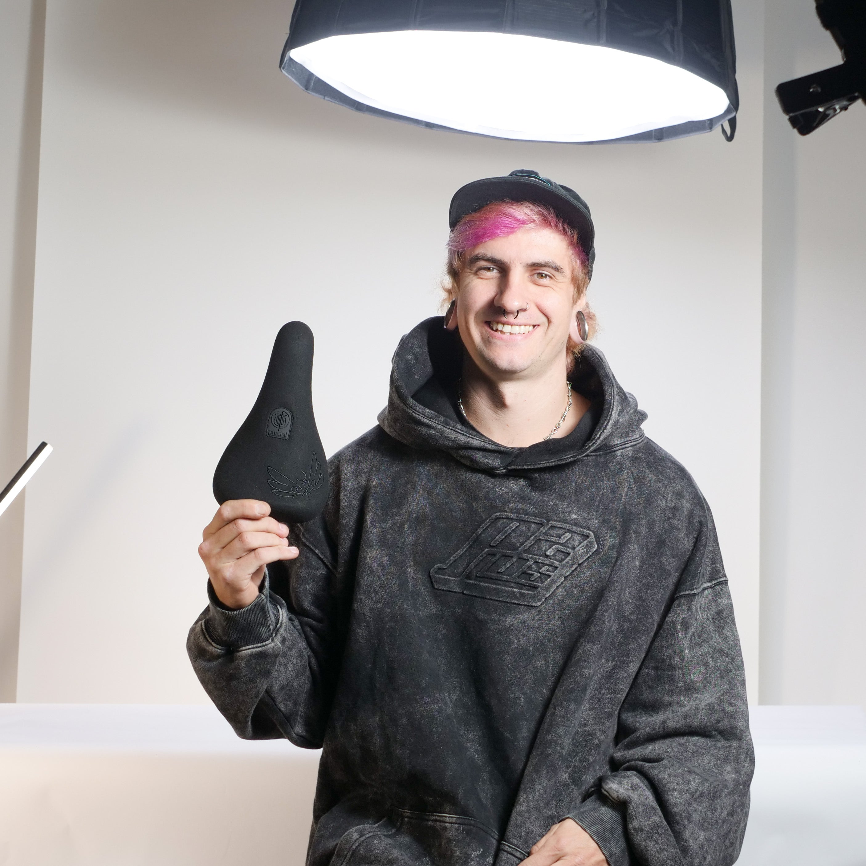 A person with pink hair, dressed in a black hoodie and cap, smiles while holding the Colony Sigil Pivotal Seat (Sean Gardner Signature) in a well-lit studio.