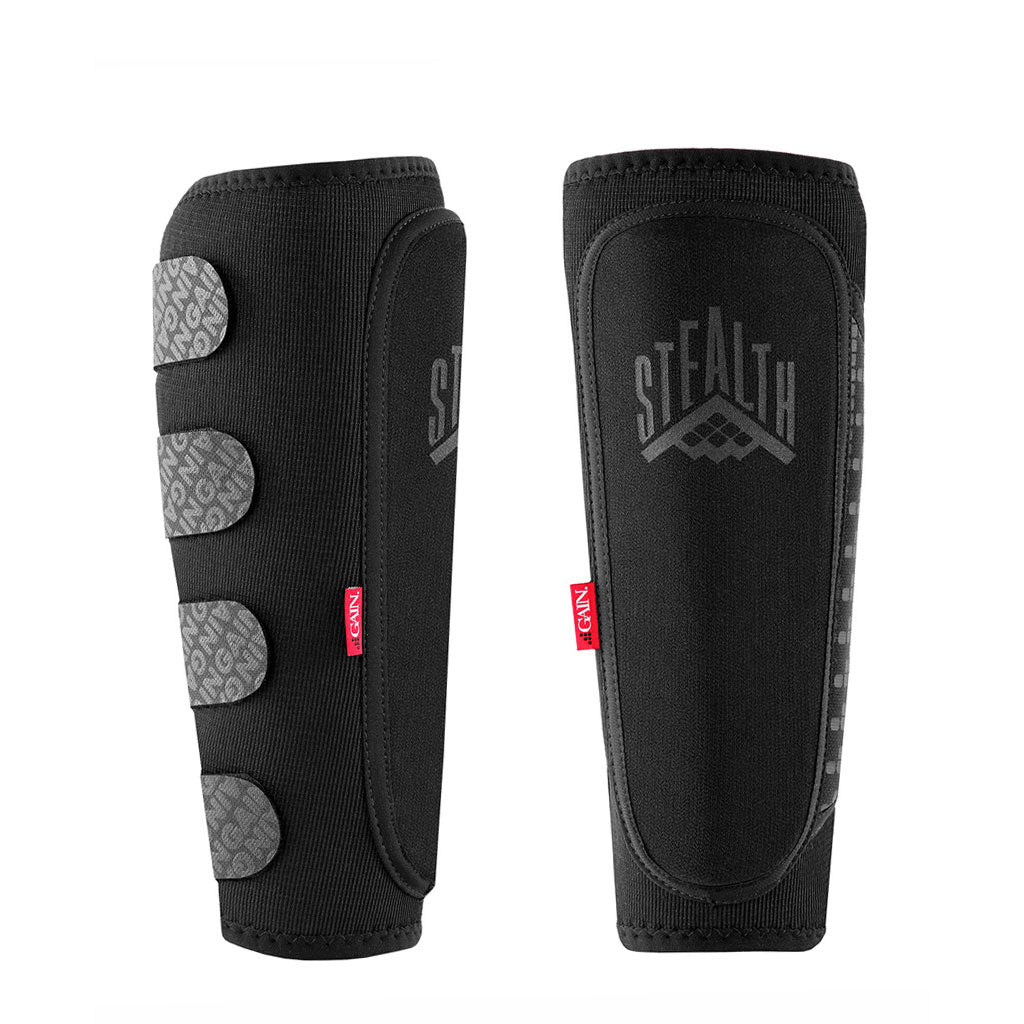 A pair of GAIN STEALTH Shin Guards in black, showcasing the gray "Stealth" logo and patterned straps on one side, crafted from DuraTec neoprene for enhanced comfort and resilience, displayed against a white background.