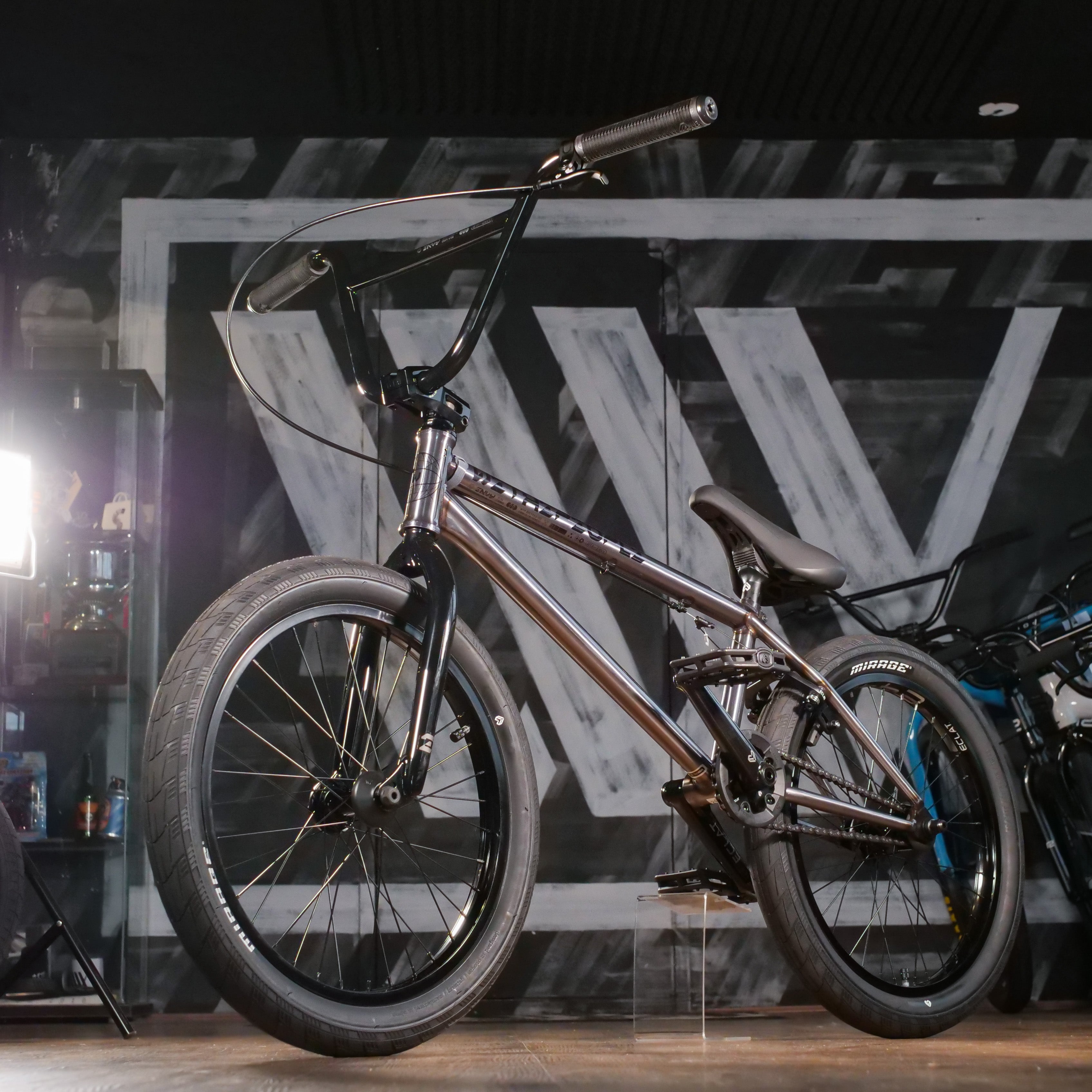 A Wethepeople Envy 20 Inch chrome BMX bike with thick tires is displayed indoors in front of a large black-and-white wall mural featuring a bold "W".