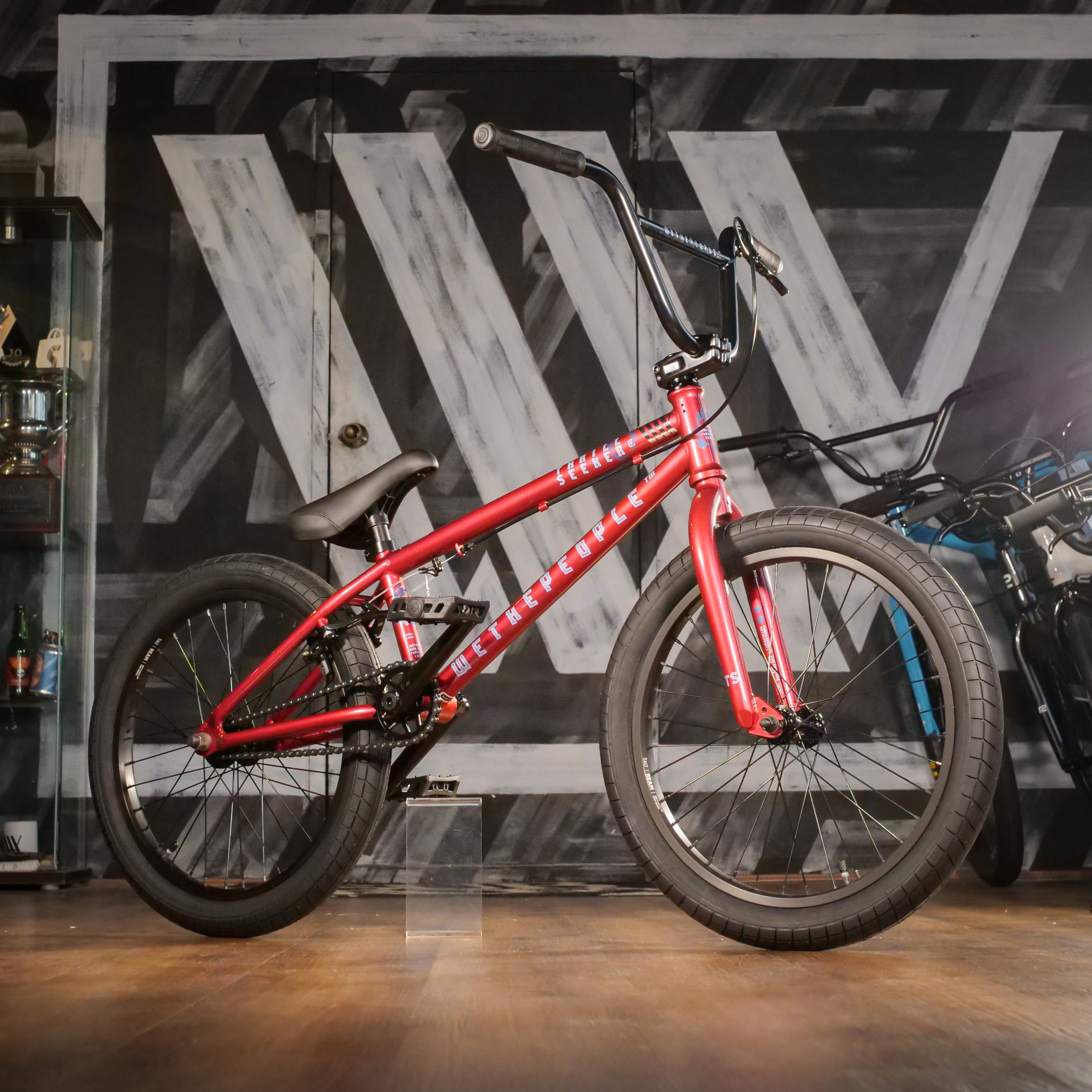 The Wethepeople Thrillseeker 20 Inch Bike in red, featuring black handlebars and tires, is displayed indoors on a wooden floor with a large "W" painted on the wall in the background.