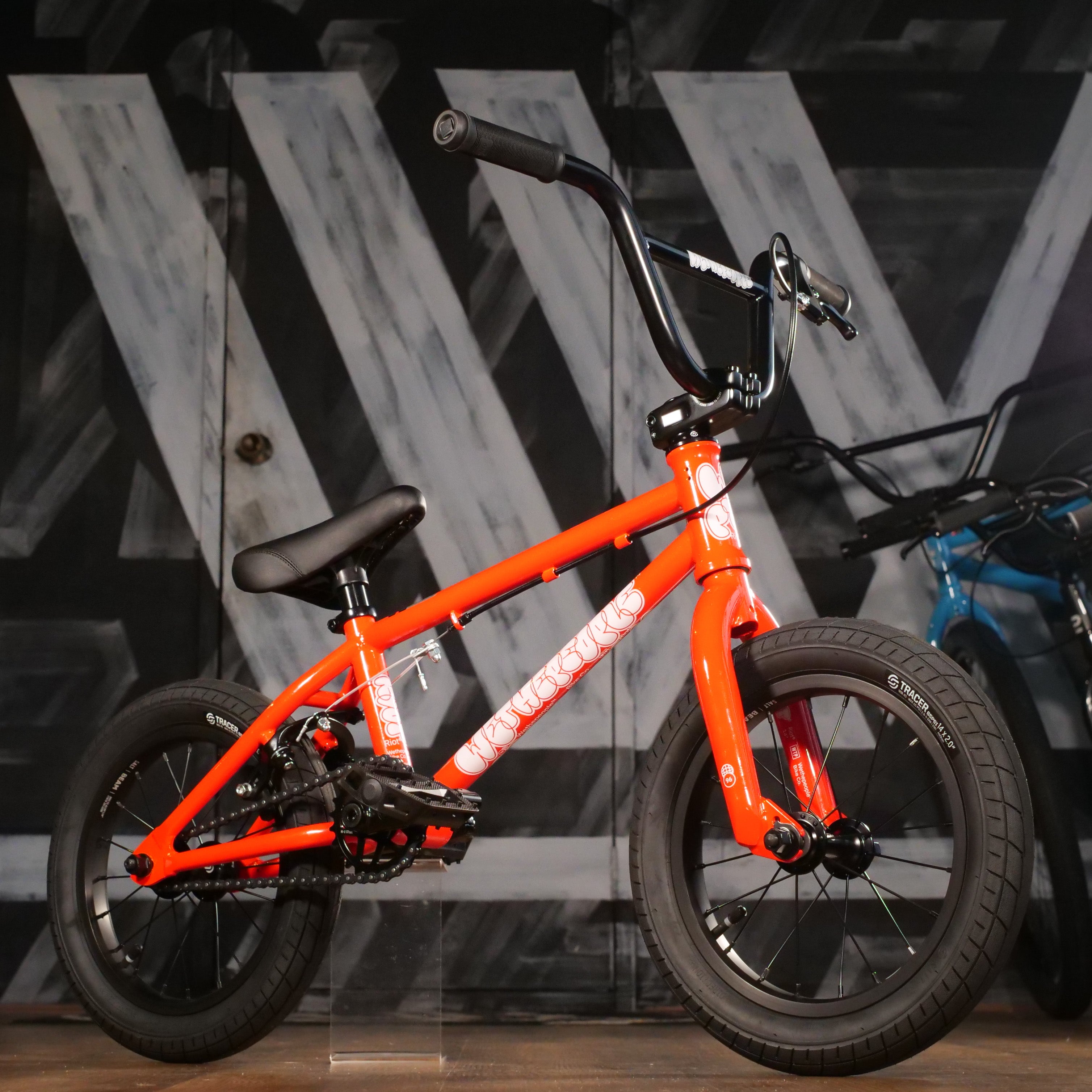 The Wethepeople Riot 14 Inch Bike is a small, bright orange kids' BMX with black tires and handlebars, shown indoors against a black and white patterned wall.