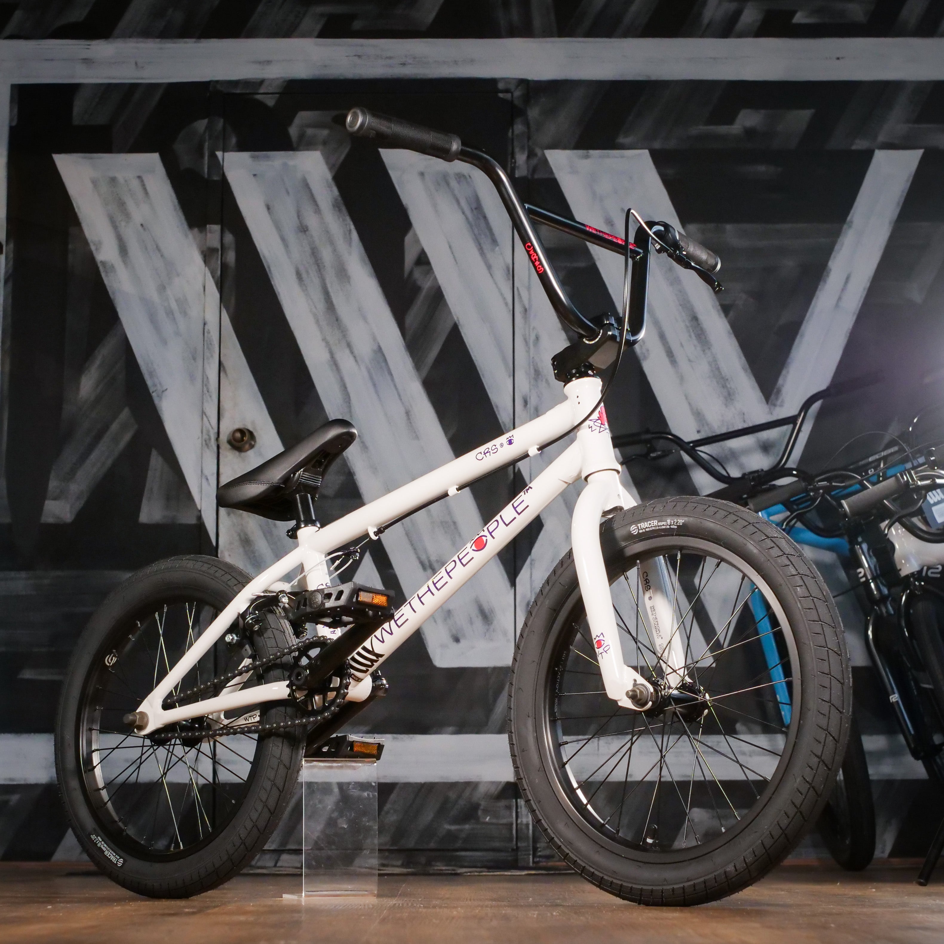 A Wethepeople CRS 18 Inch Bike in white with black tires and handlebars is showcased indoors against a dark, patterned wall.