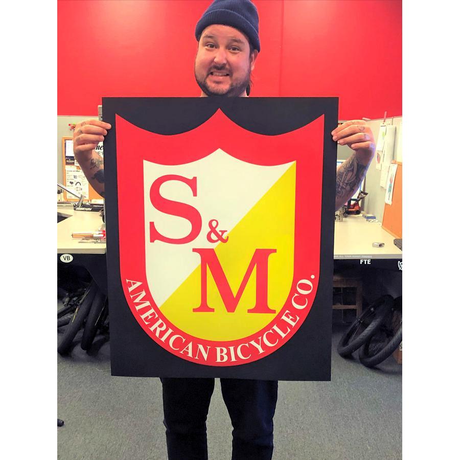 A man holds an S&M Shield Banner featuring the American Bicycle Co. logo in an office, ready to showcase your love for your favorite brand.