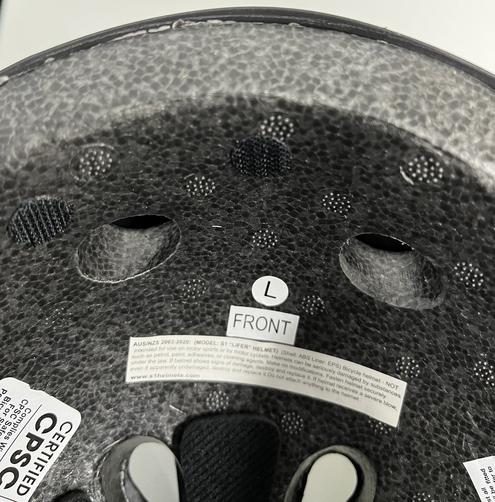 The inside of a bicycle helmet displaying texture, ventilation holes, label 'FRONT', size 'L', and safety certification stickers, equipped with a handy S-One Lifer (12 Pack) Velcro Kit Round Dots for easy adjustments.