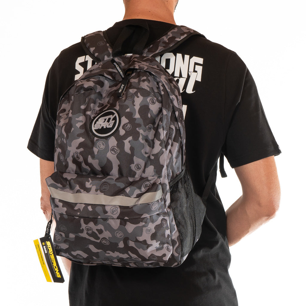 A person is wearing the Staystrong Backpack Chevron V3, featuring a gray and black camouflage design, a front pocket, mesh side pocket, and adjustable shoulder straps. They are also dressed in a black t-shirt with white lettering.