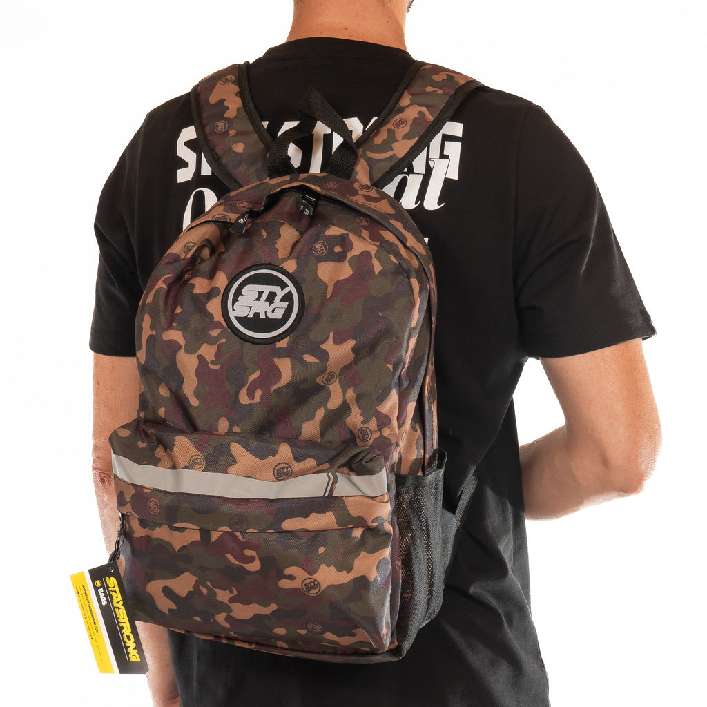 A person wearing a black T-shirt carries a Staystrong Backpack Chevron V3 featuring a camouflage pattern, mesh side pockets, adjustable shoulder straps, and a yellow tag attached.