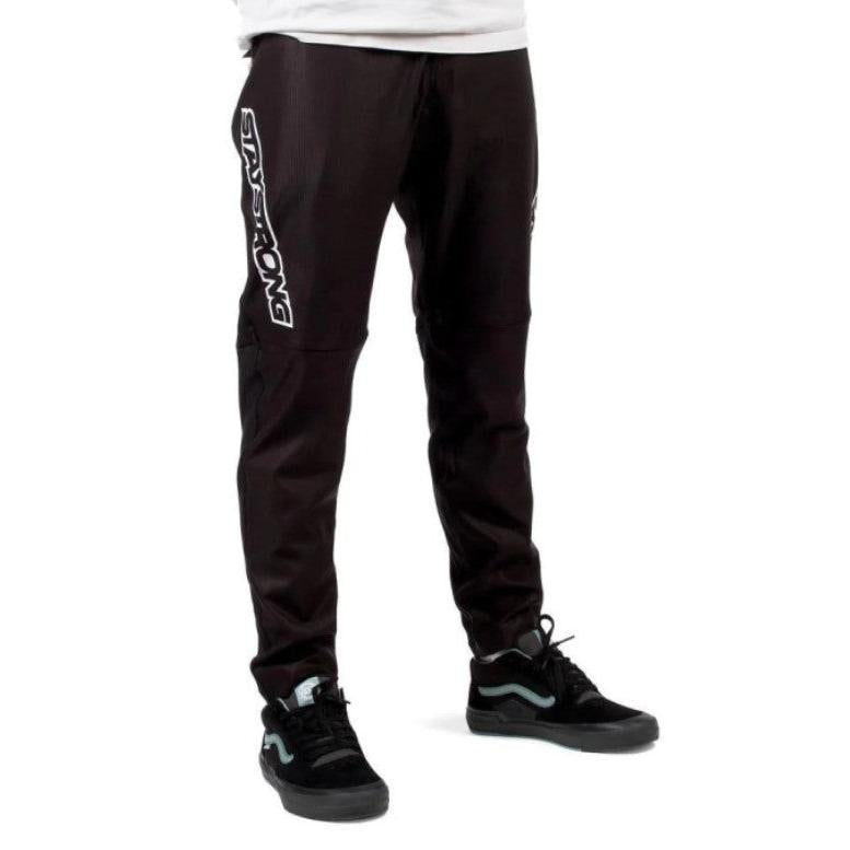 A person wearing Staystrong V3 Race Pants, featuring “Stay Strong” printed on one leg, paired with black Vans sneakers and a white shirt.