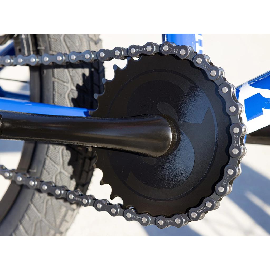 Close-up of the black front chainring and chain on a Sunday Blueprint 16 Inch Bike, with part of the blue frame and rear tire visible in the background.