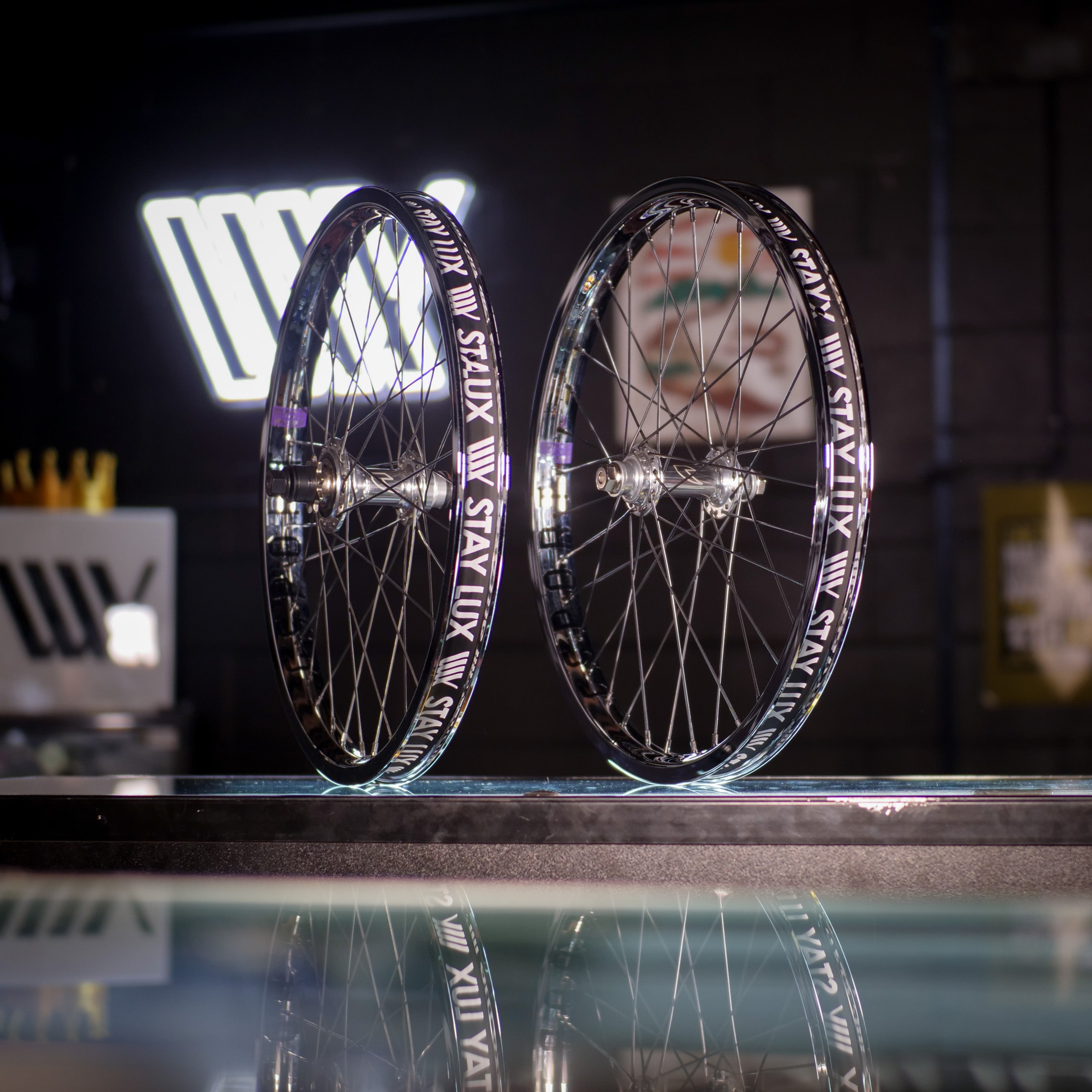 Two Tall Order X Birdcage Custom Wheelsets stand upright on a workshop counter, with reflective accents and blurred shop signage in the background for an authentic vibe.