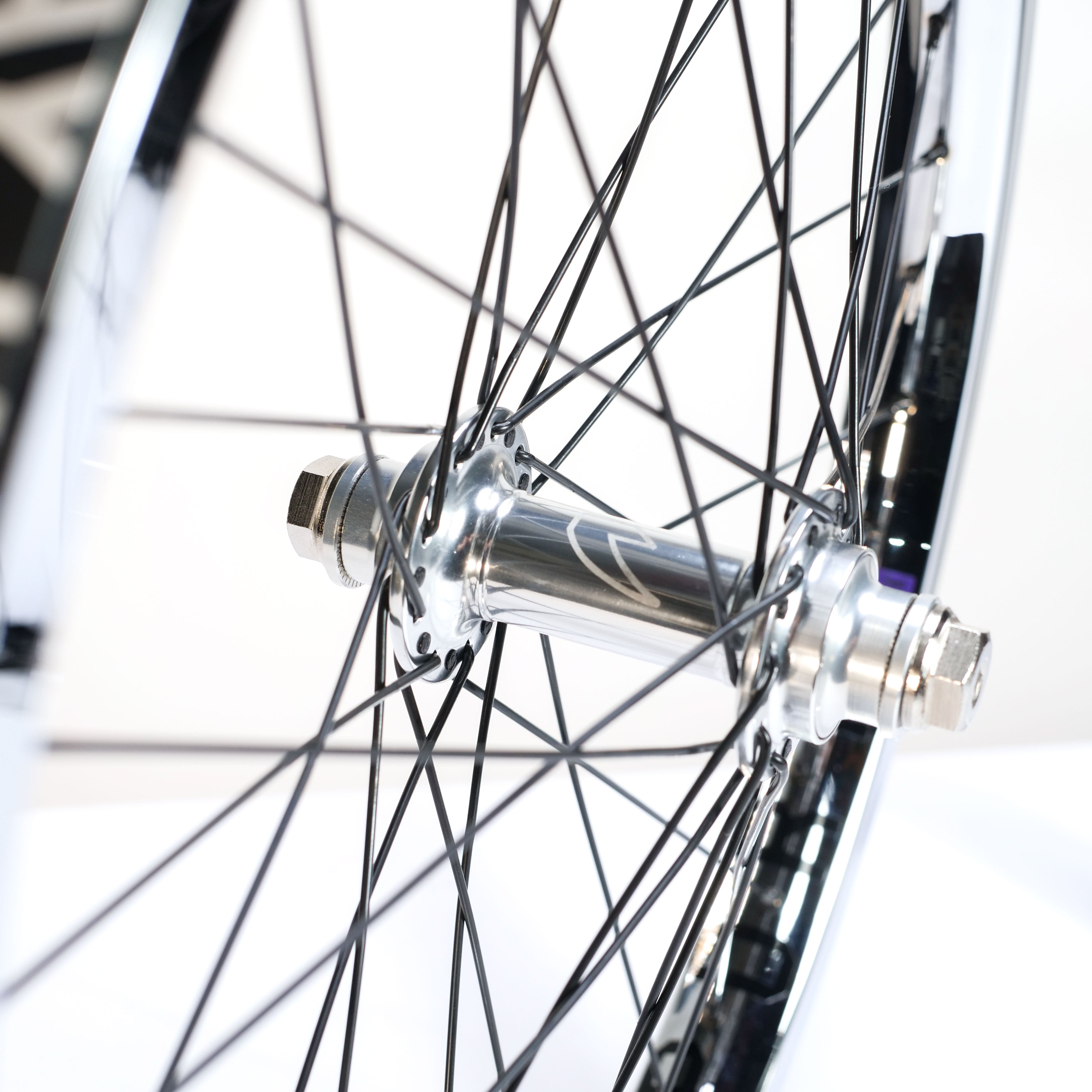 Close-up of a bicycle wheel with a silver hub, black spokes, and axle with nuts against a light background—showcasing the quality details of the Tall Order X Birdcage Custom Wheelset.