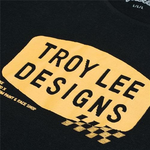 A close-up of the TLD 25.1 Stamp SS T-Shirt reveals a stylish black tee with a yellow rectangular design showcasing "Troy Lee Designs" in bold. Smaller text and a checkered pattern below add flair, embodying true TLD pride.