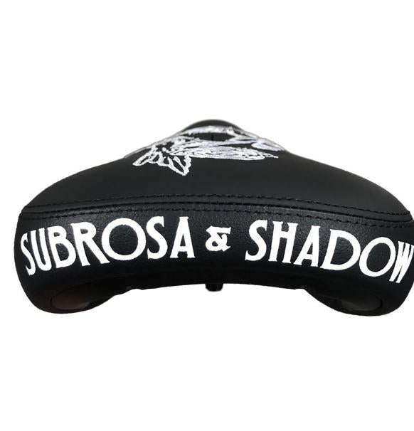 The Subrosa x Shadow Rose Crow Pivotal Seat features bold white "SUBROSA & SHADOW" lettering on the back and a unique top design—ideal for riders seeking a quality, compatible seat with signature style.