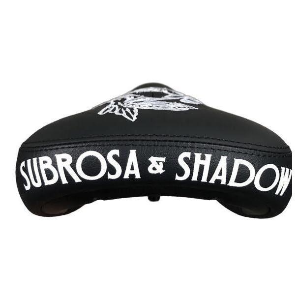 The Subrosa x Shadow Rose Crow Pivotal Seat features bold white "SUBROSA & SHADOW" lettering on the back and a unique top design—ideal for riders seeking a quality, compatible seat with signature style.