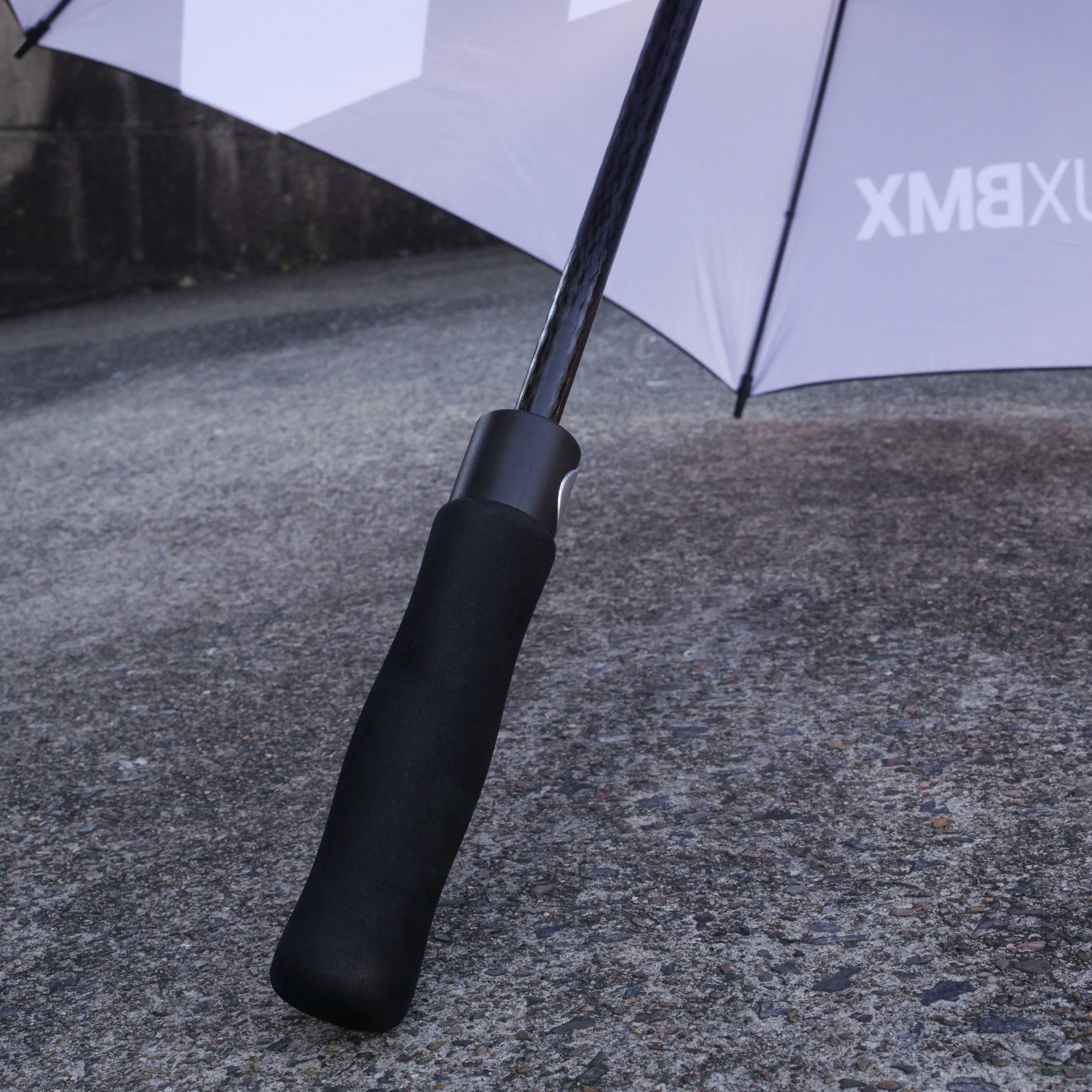 Close-up of a LUXBMX Race Track Umbrella handle on a textured outdoor surface, with the premium carbon pole umbrella open in the background.