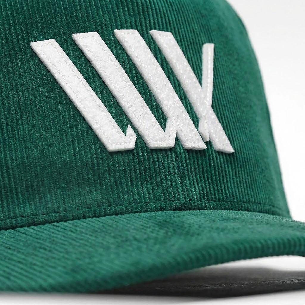 Close-up of the LUXBMX Outfield Cap in Bottle Green, a corduroy baseball cap with an embroidered LUX logo and overlapping white geometric lines on the front panel.