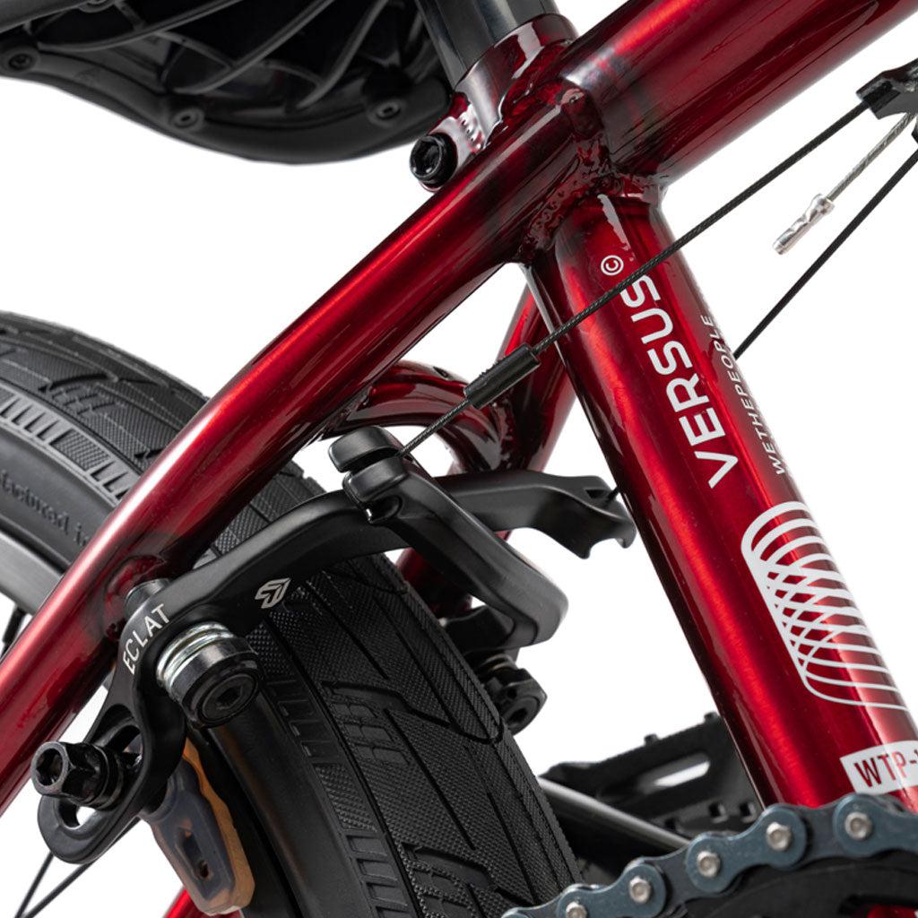 Close-up of a Wethepeople Versus 20 Inch Bike, showing its red chromoly frame labeled "VERSUS" with a white spiral graphic, rear tire, Eclat brake assembly, cables, and part of the seat.
