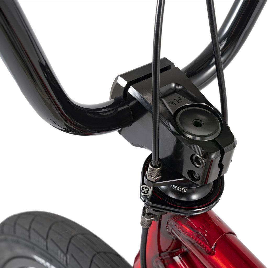 Close-up of a black BMX handlebar and stem with brake cables, plus part of the red Chromoly frame and front tire of the Wethepeople Versus 20 Inch Bike.