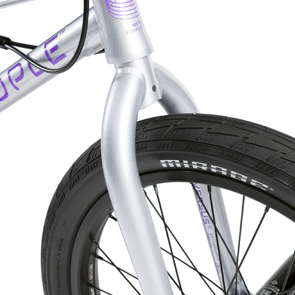 Close-up of a Wethepeople Versus 20 Inch Bike’s silver Chromoly fork and front wheel, "MIRAGE" text on the tire, purple branding on the frame, and contrasting black spokes.