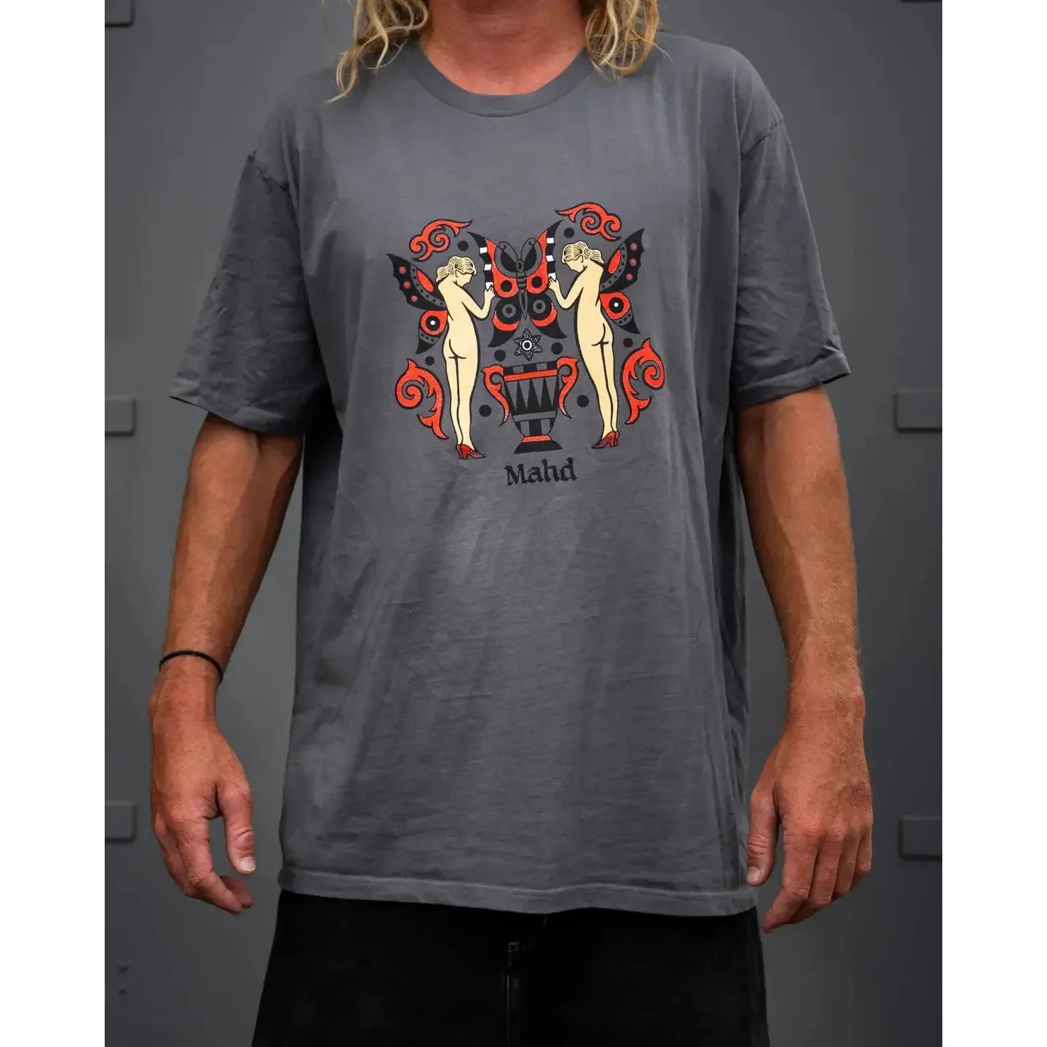 A person wears the Mahd Wesley Tee in gray, showcasing a traditional tattoo-style graphic with two nude female figures, red and black wing shapes, and "Mahd" printed below—design inspired by Cape Town’s dynamic art scene.