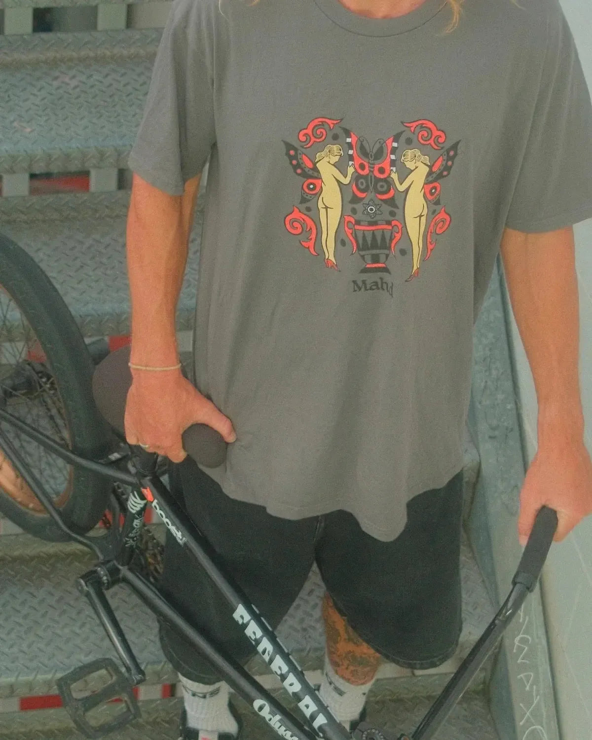 A person stands on a metal staircase in Cape Town, holding a black BMX bike and wearing the Mahd Wesley Tee with a vibrant traditional tattoo-inspired graphic, paired with black shorts.