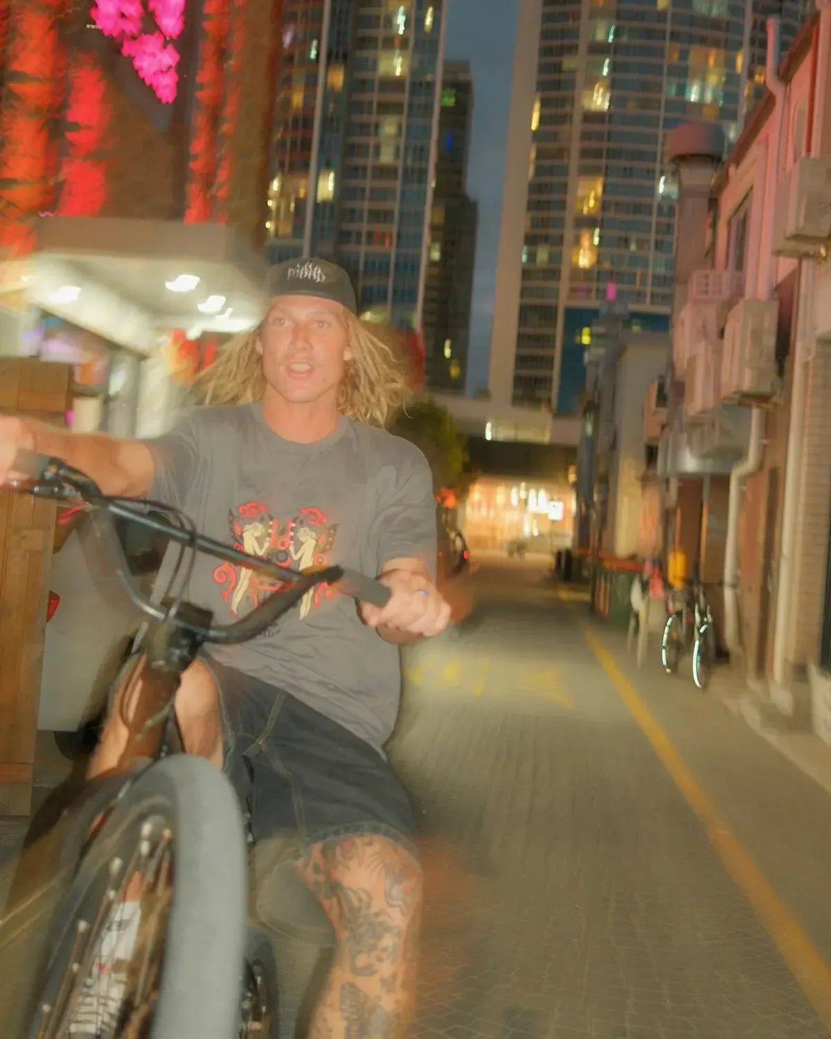 Wearing the Mahd Wesley Tee, a person with long blond hair and traditional tattoos rides a bike on one wheel down a Cape Town street at dusk, tall buildings rising in the background.