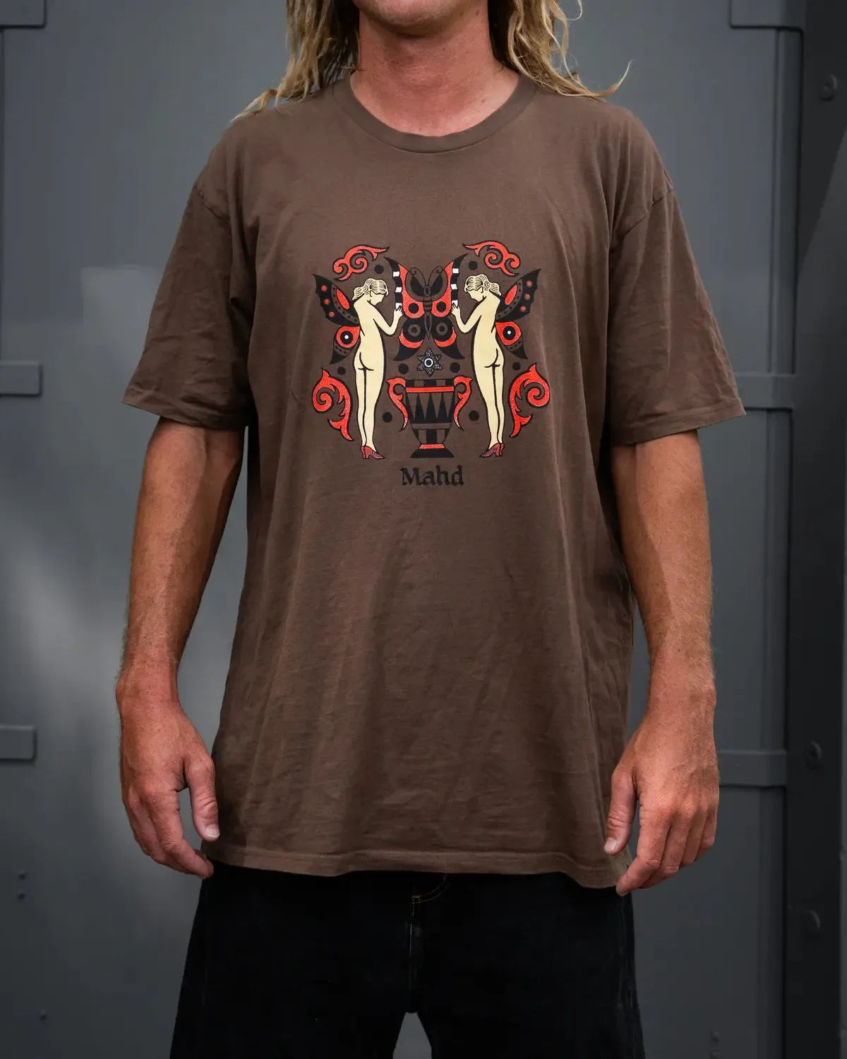 Someone wears the Mahd Wesley Tee, a brown shirt featuring an artistic graphic of two nude figures and a decorative urn in traditional tattoo style, with "Mahd" below the design.