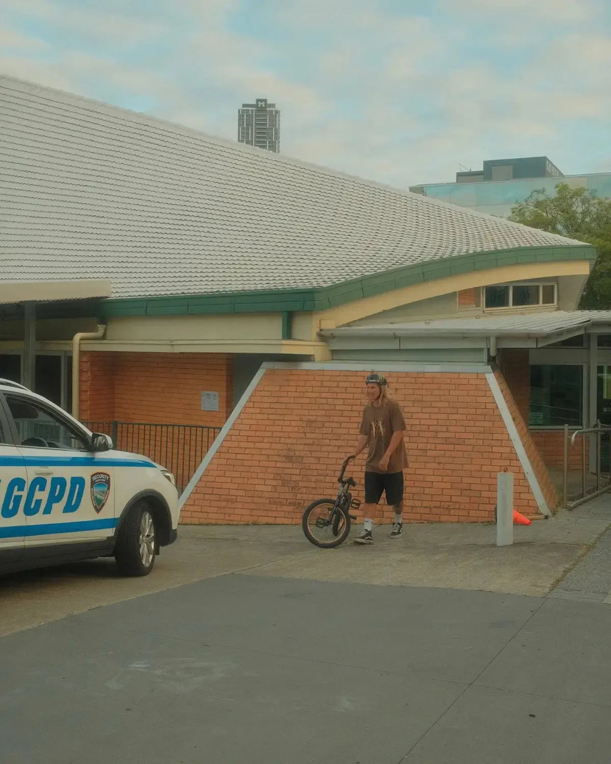 In Cape Town, a BMX rider stands by a brick building near a GCPD police car, wearing the Mahd Wesley Tee featuring traditional tattoo style graphics.