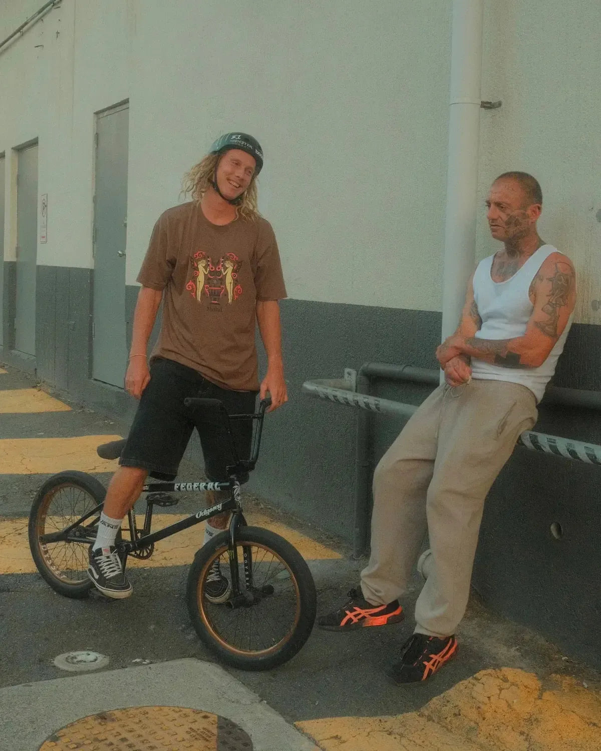Two men outside a building in Cape Town: one stands with a BMX bike, wearing a helmet and casual clothes featuring the Mahd Wesley Tee; the other leans against a railing in a tank top and sweatpants.