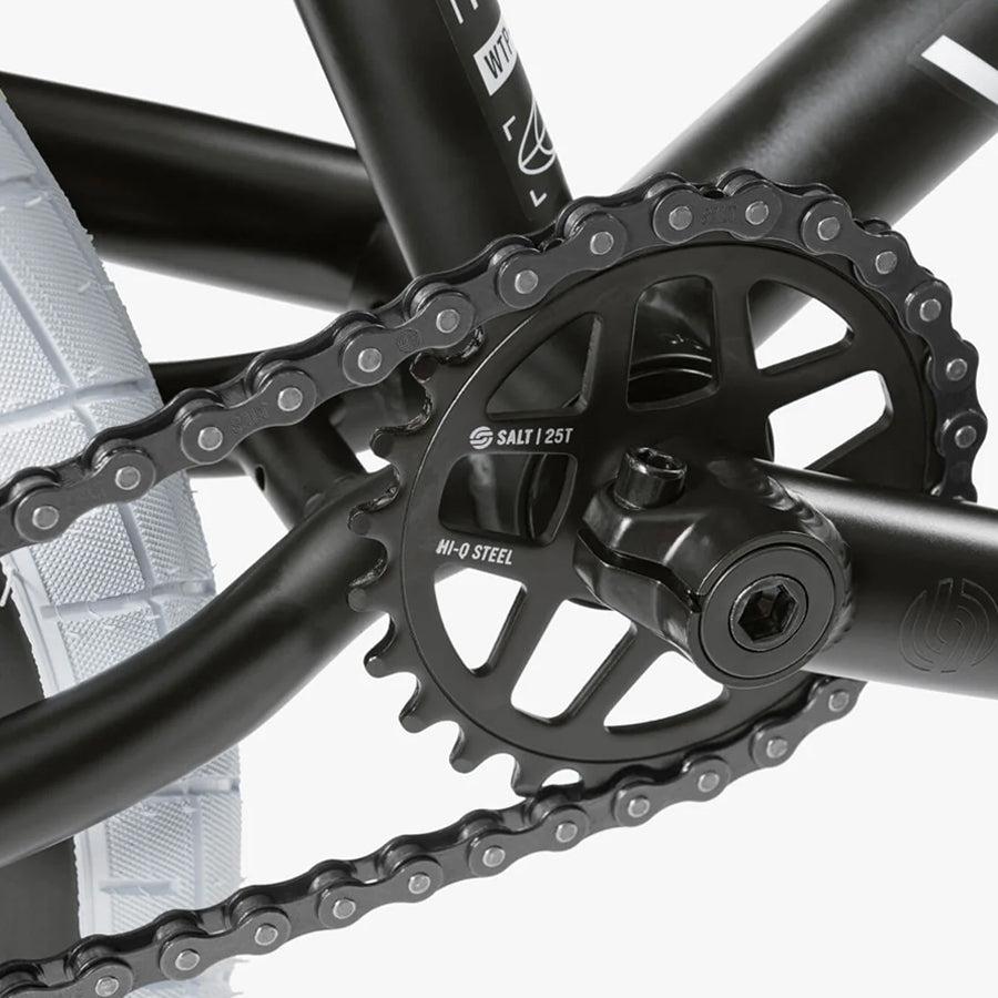 Close-up of a black bicycle chainring and chain with "Salt/25T Hi-Q Steel" engraved. The rear wheel and tire of the Wethepeople Nova 20 Inch Bike are visible, highlighting its impressive build with robust Salt Rookie Cranks.