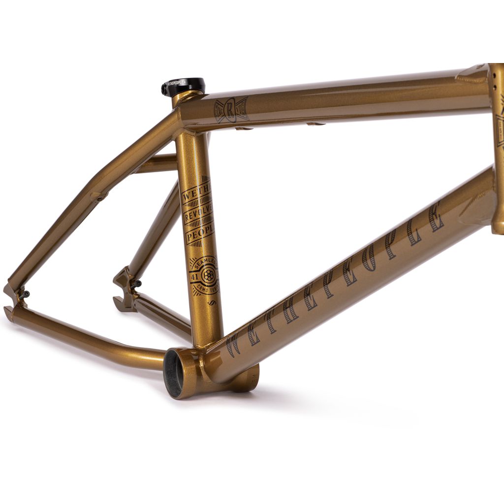 The Wethepeople Revolver Frame (2022) is a gold BMX frame constructed from seamless 4130 crmo tubing, adorned with black text and designs. It is showcased at an angle against a white background and includes a lifetime guarantee for durability.