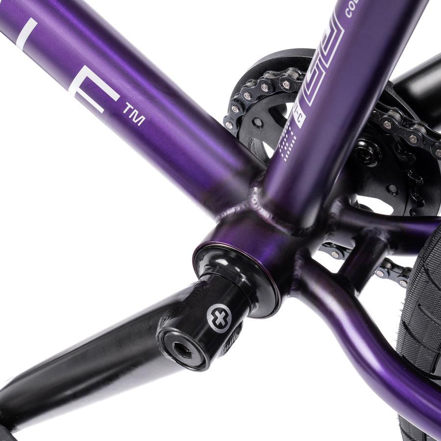 Close-up of the Wethepeople Trust 20 Inch Freecoaster/Hybrid Bike's purple frame's bottom bracket area, highlighting the precise battleship geometry in the crank arm and chainring design.